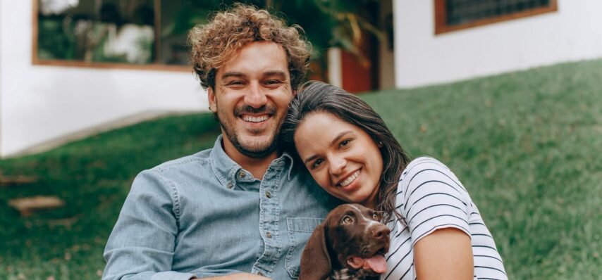 A joyful couple sitting on grass with their dog, enjoying a sunny day outdoors.